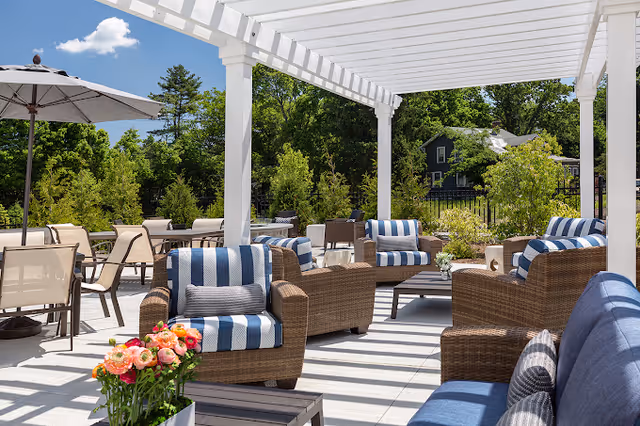 Outdoor patio with wicker lounge chairs and striped cushions under a white pergola overlooking landscaped greenery.