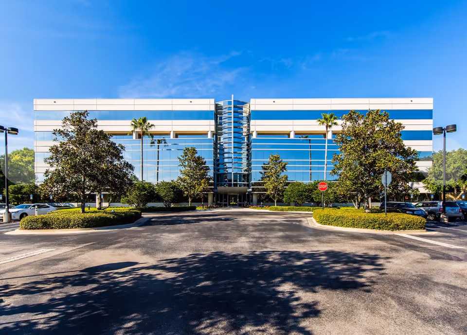 Modern glass-front building with palm trees, driveway, and parking in a sunny lot.