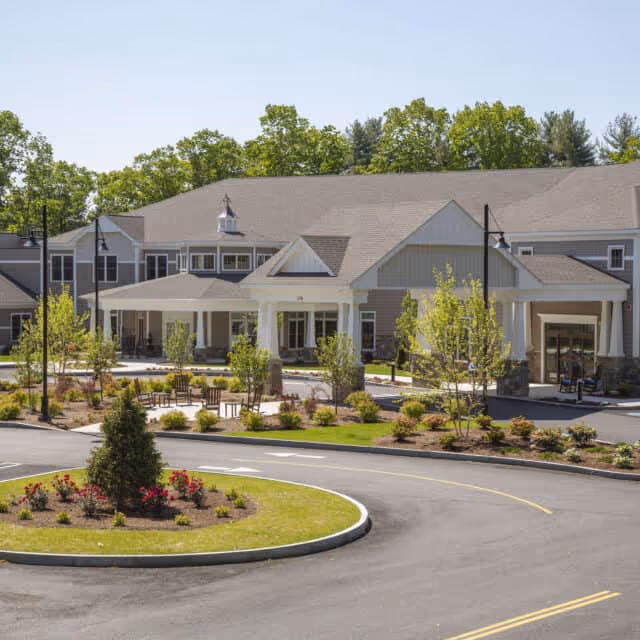 Front exterior view of a senior living facility named Cornerstone at Hampton, featuring a large building with a covered entrance, landscaped garden beds with shrubs and flowers, a circular driveway, and trees in the background under a clear sky.