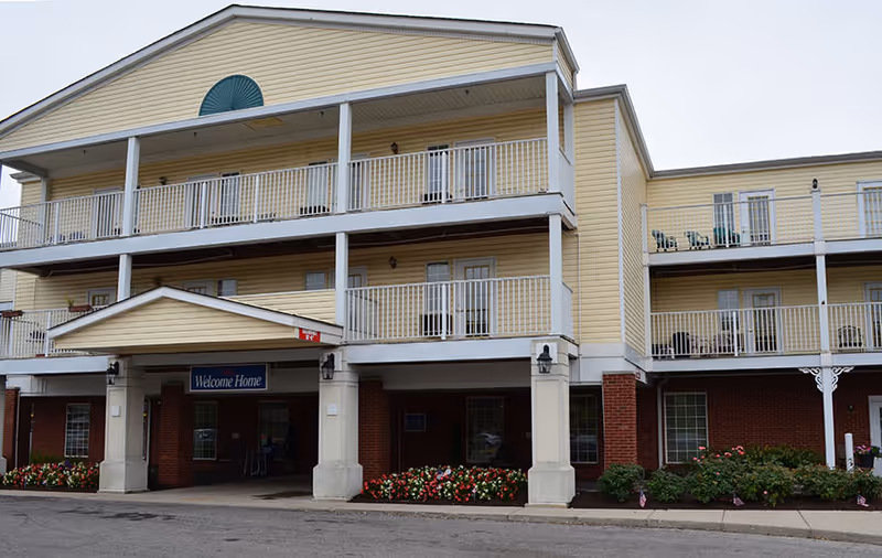 Front exterior of a three-story yellow-and-brick senior living building with balconies and a covered entrance sign reading 'Welcome Home'.