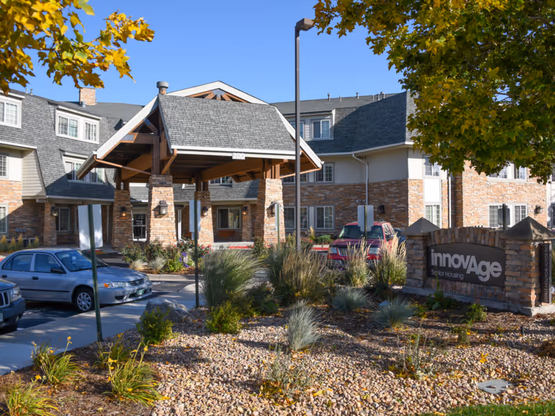 Front entrance of a senior living facility with a covered porte-cochere, landscaped rock garden, parked cars, and a sign reading "innovAge Senior Housing".