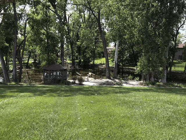 A peaceful outdoor area with a well-maintained green lawn, tall trees, a small wooden gazebo, and a wooden dock extending over a pond or small body of water.