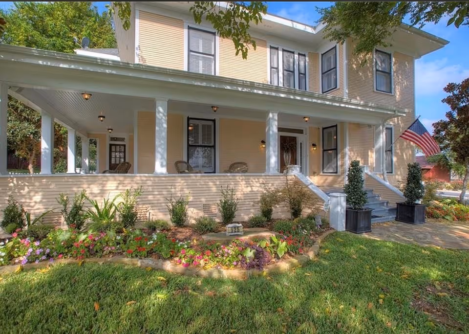 A yellow two-story building with a wide front porch, white columns, and a flower garden.