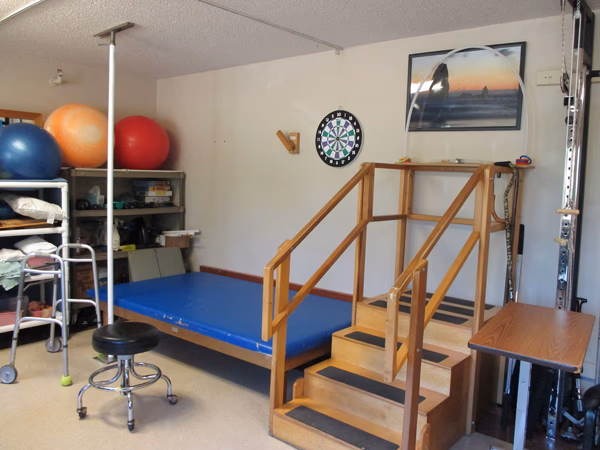 Room with wooden gait-training stairs, blue therapy mat, large exercise balls, a walker, and a rolling stool.
