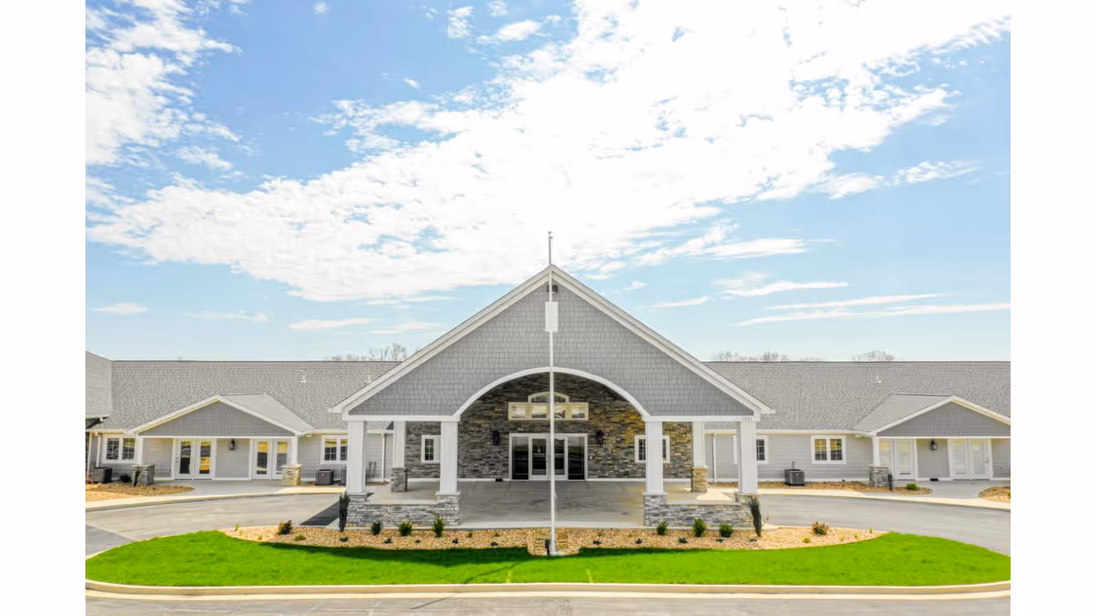 Front exterior view of Cedarhurst Senior Living of West Plains building with a large covered entrance, stone and gray siding facade, and a well-maintained lawn under a partly cloudy sky.