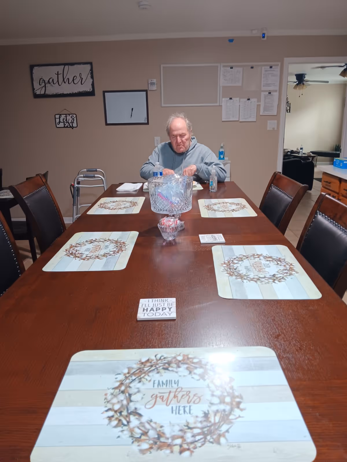 An elderly man sits at a long wooden dining table in a senior living facility.