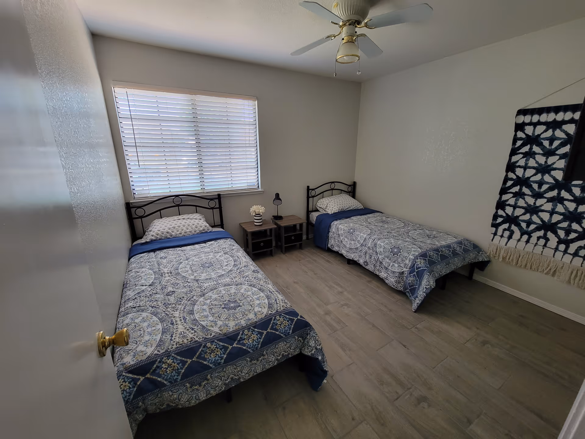 Two twin beds with blue patterned bedding sit in a bedroom with a window and fan.