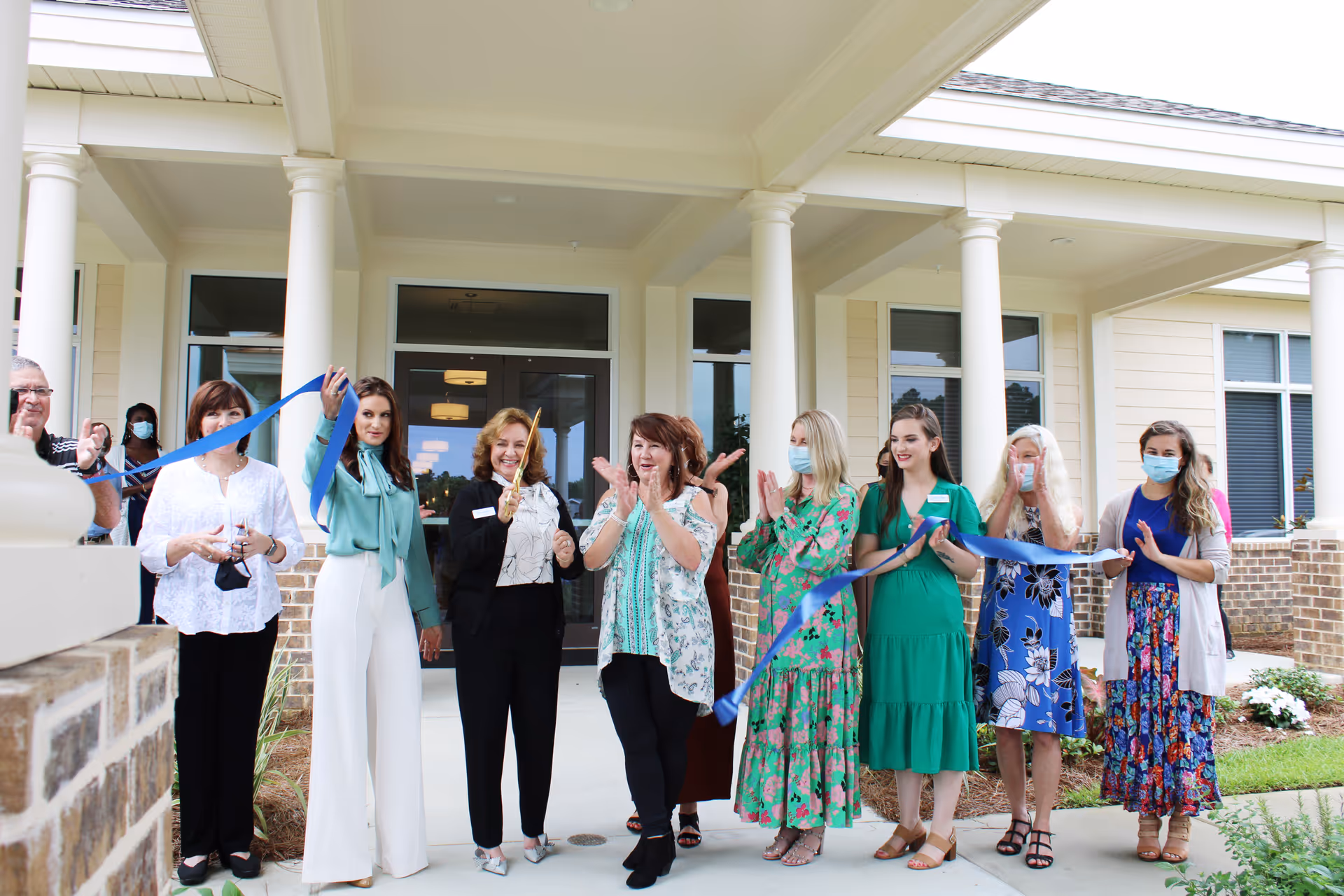 A group of people standing in front of a building entrance participating in a ribbon-cutting ceremony. Several women are holding a blue ribbon, and one woman in the center is holding large scissors. Some individuals are wearing face masks. The building has white columns and a covered porch.