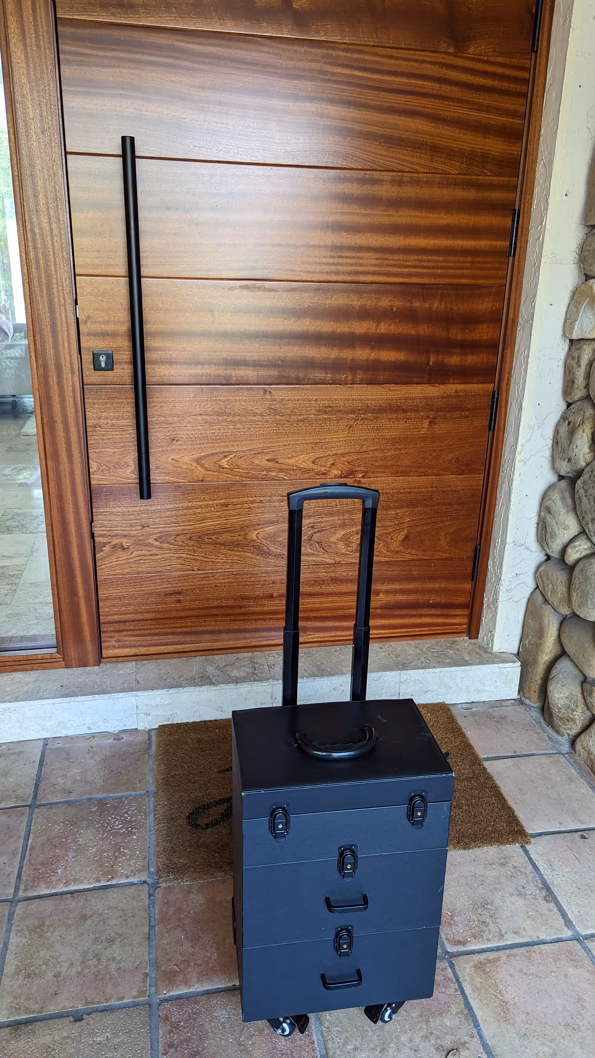 Tall wooden front door with black handle beside rolling gray cabinet on a tiled entry.