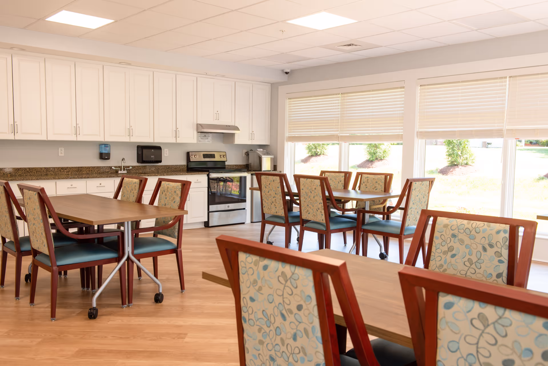 Bright communal dining area with wooden tables and patterned chairs facing a kitchenette and large windows.