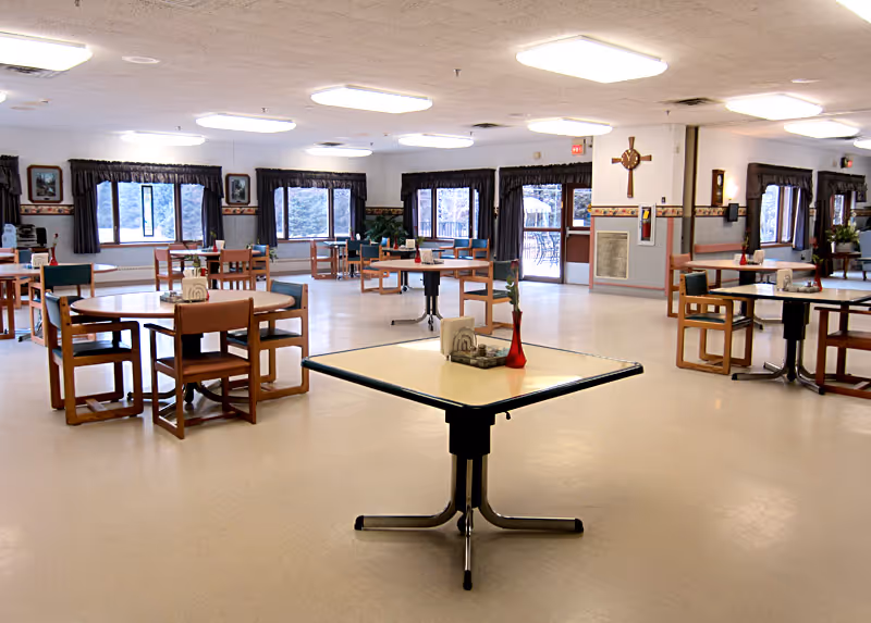 A spacious dining room with multiple tables and chairs arranged neatly. Each table has a small centerpiece and napkin holder. Large windows with dark curtains allow natural light to fill the room. The walls are decorated with framed pictures and a large cross is mounted on one wall. The floor is light-colored and the ceiling has several rectangular light fixtures.