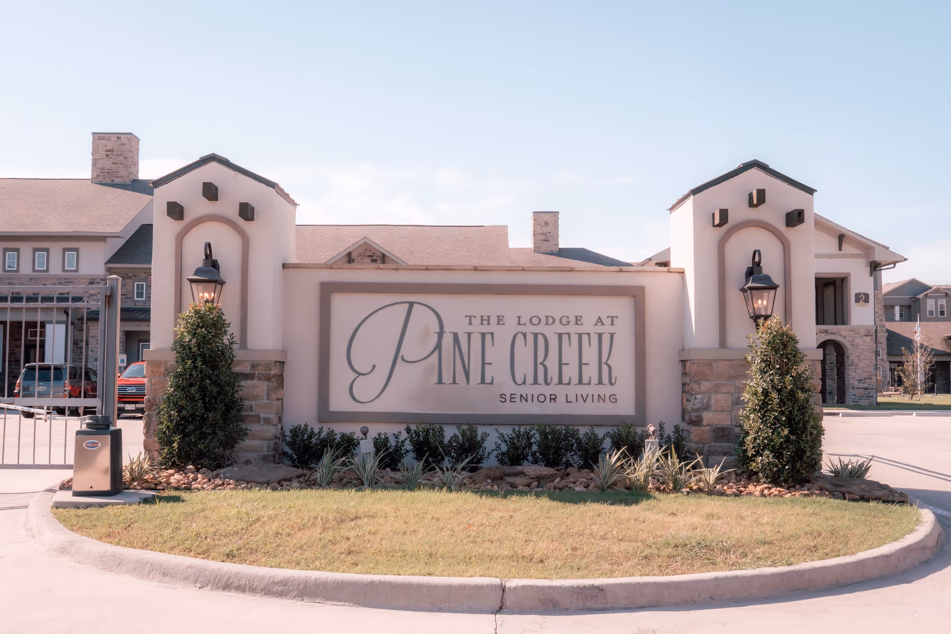 Entrance sign for The Lodge at Pine Creek Senior Living, featuring a large stone and stucco structure with two lanterns and landscaping in front. Behind the sign, parts of the senior living facility buildings and a gated driveway are visible under a clear sky.