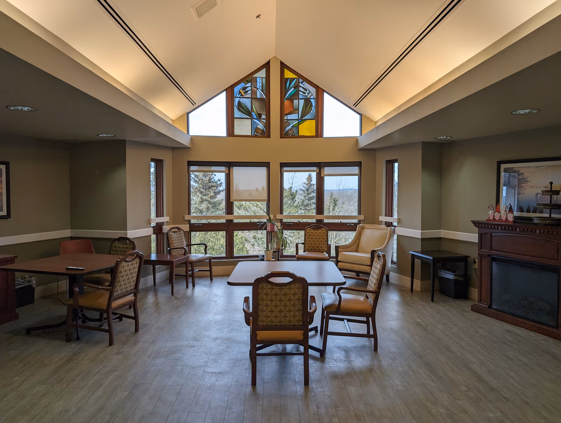 Sunlit common living area with tables, chairs, and large windows in a senior facility.