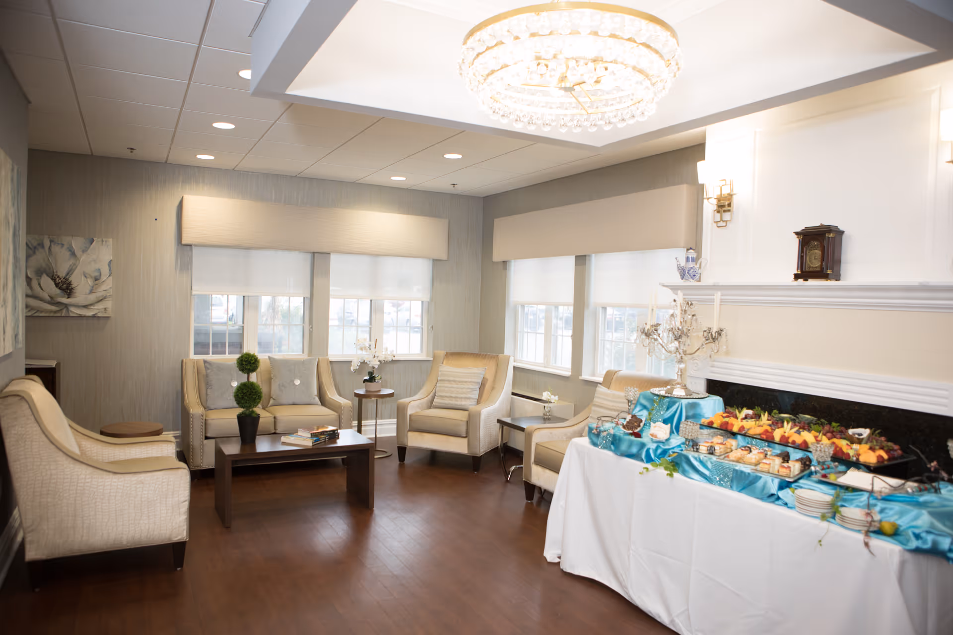 A bright and elegant living room area in CareOne at Wayne Assisted Living featuring beige upholstered chairs and sofas arranged around a wooden coffee table with a small plant and books. Large windows with beige valances allow natural light to fill the room. On the right side, a table covered with a white tablecloth and blue satin runner displays an assortment of food and desserts. A crystal chandelier hangs from the ceiling, and a decorative clock sits on the mantel above a fireplace.