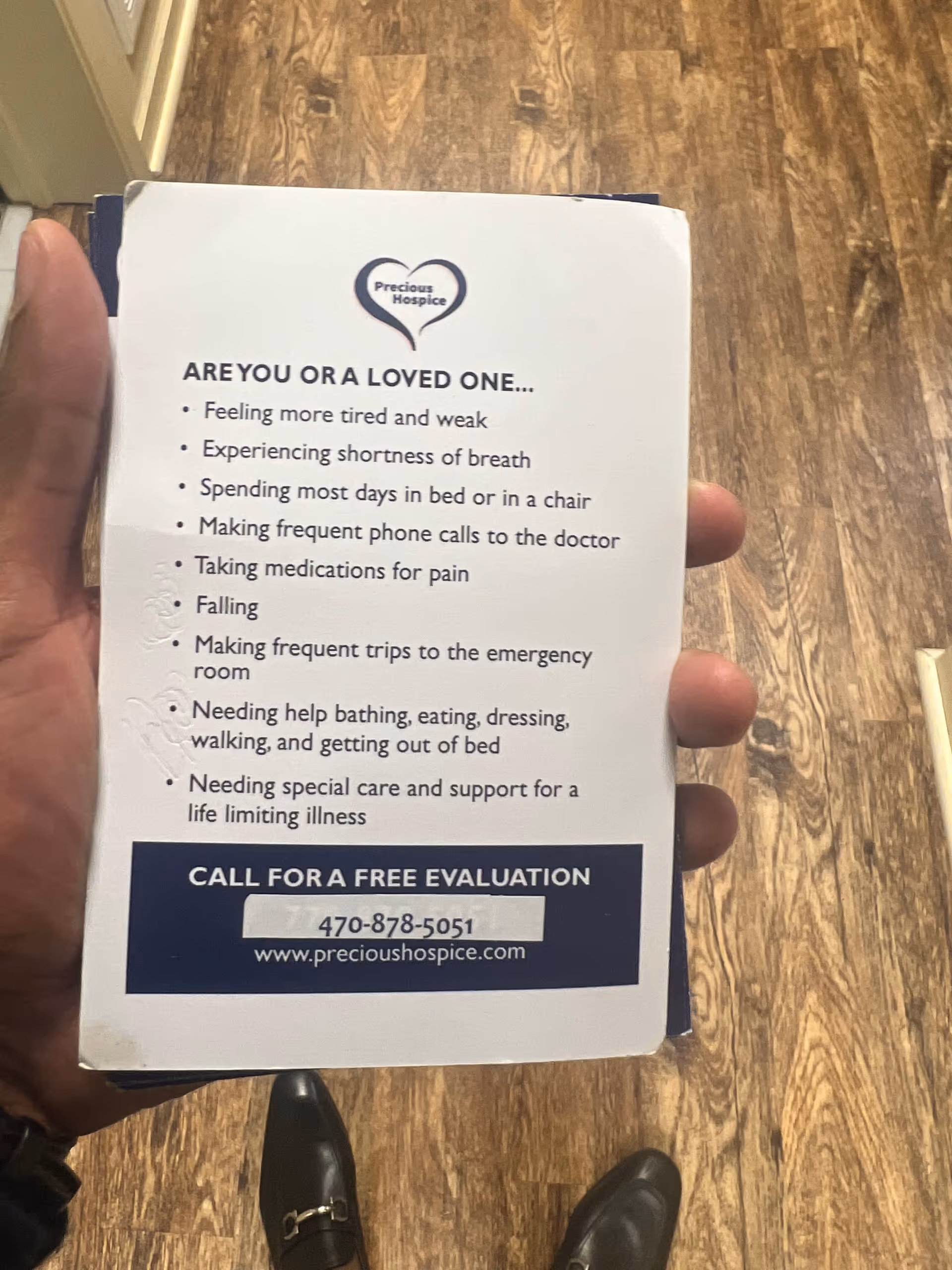 Hand holds a Precious Hospice brochure in a hallway with wood flooring.