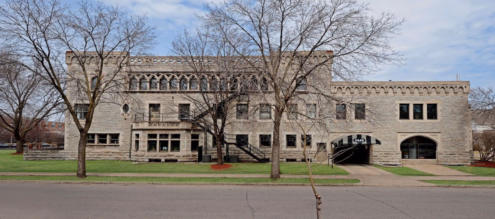 Stone two-story building with arched windows and a driveway, trees lining a grassy front.