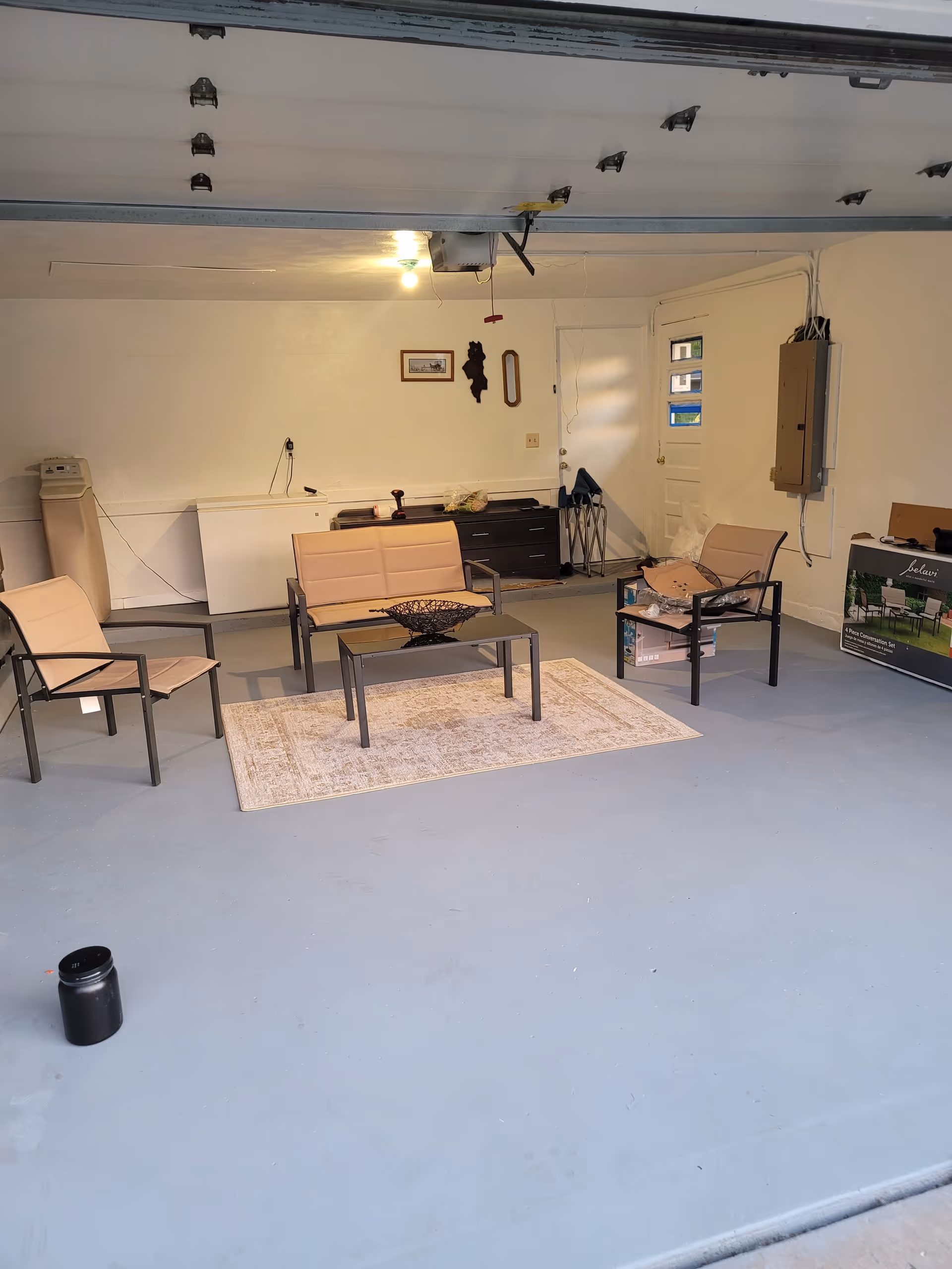 An indoor space resembling a garage with an open garage door. The room has a seating area with two chairs and a loveseat around a small coffee table on a rug. There is a cabinet and some miscellaneous items against the back wall, including a folded chair and a box. The floor is painted gray and the walls are white.