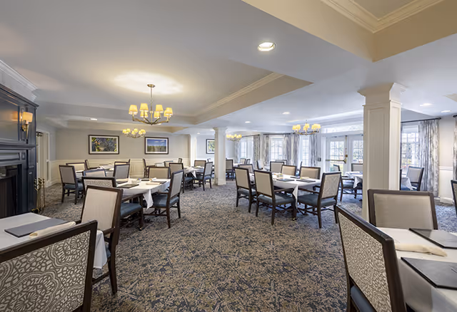 A spacious dining room with multiple tables covered with white tablecloths and surrounded by cushioned chairs. The room features carpeted flooring, several chandeliers, large windows with curtains allowing natural light, and framed artwork on the walls.