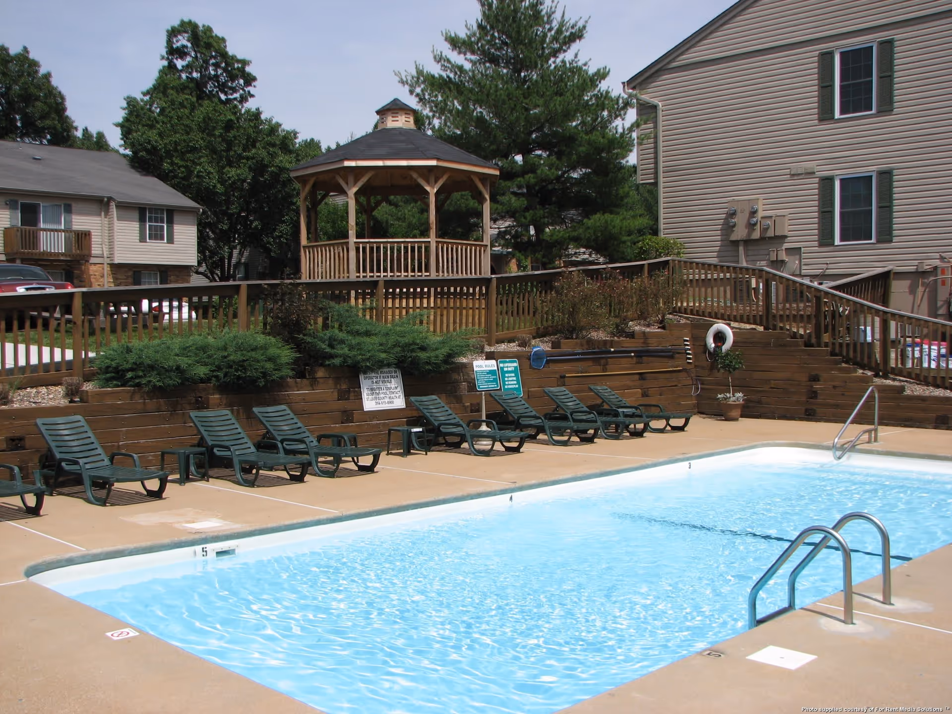 Outdoor swimming pool with clear blue water surrounded by a concrete deck. Several green lounge chairs are lined up along one side of the pool. Behind the chairs is a wooden fence with bushes and a wooden gazebo. Residential buildings and trees are visible in the background under a clear sky.