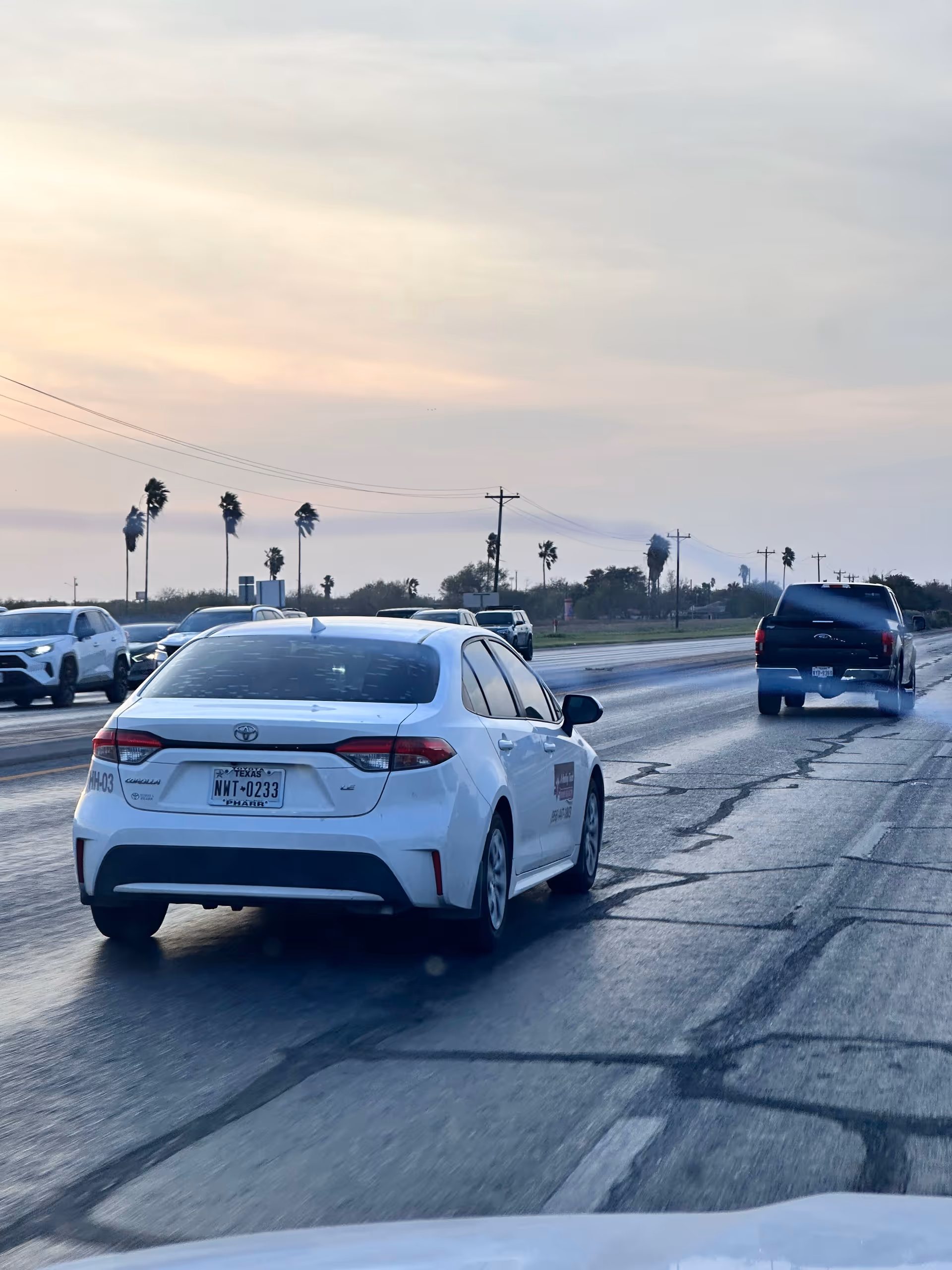 White sedan on a wet highway with palm trees and other vehicles at dusk.