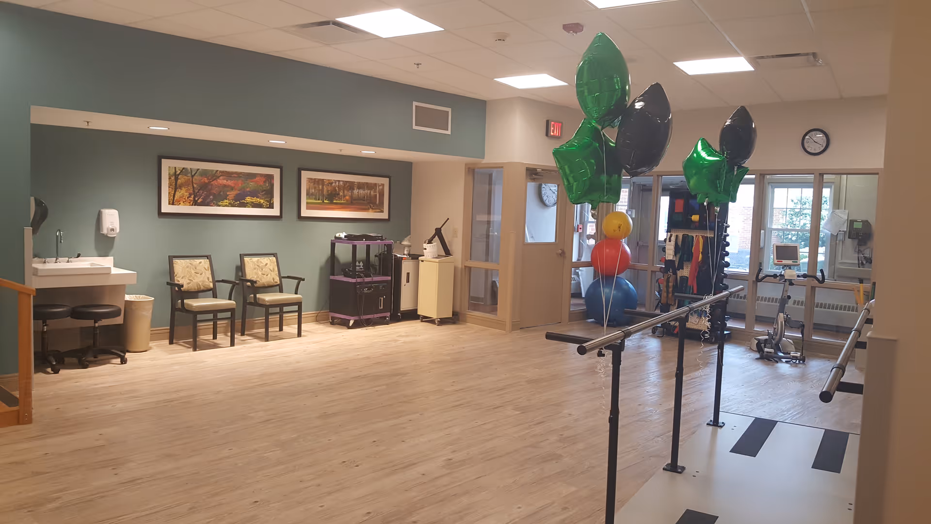 Spacious rehabilitation therapy room with parallel walking bars, exercise equipment, chairs and decorative balloons.