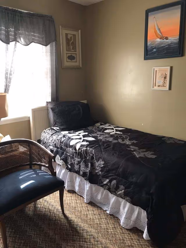 Small bedroom with a single bed, dark floral comforter, chair, window, and wall art.