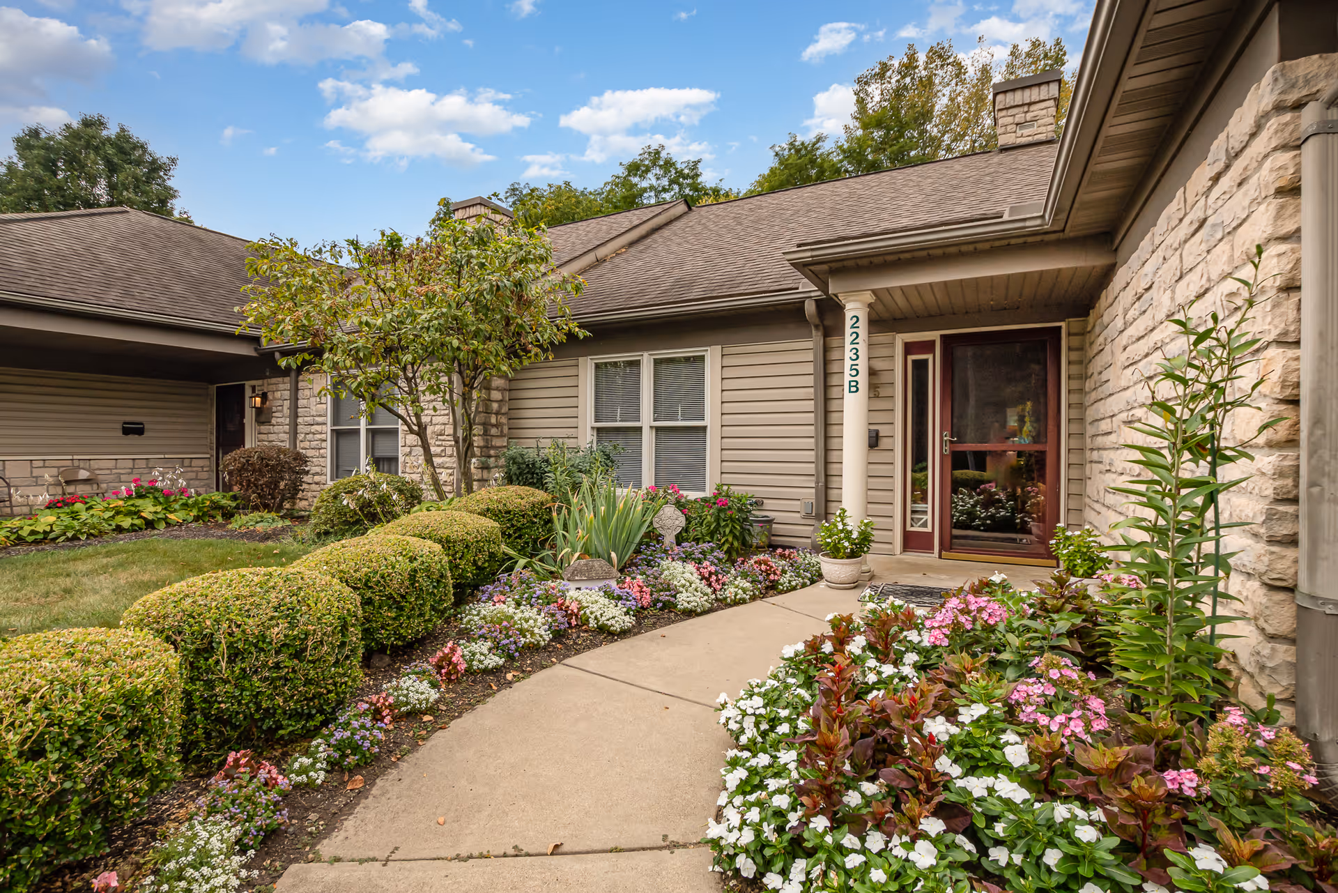 Exterior view of a single-story residential unit at Wesley Ridge with beige siding and stone accents. The entrance features a red door with glass panels, a white column, and the unit number 2235B. The walkway is bordered by neatly trimmed bushes and colorful flower beds under a partly cloudy sky.