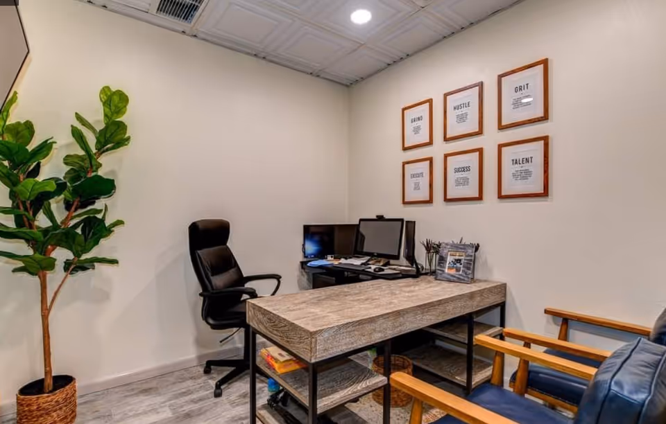 Small office with desk, computer monitors, plant, and framed motivational quotes on the wall.
