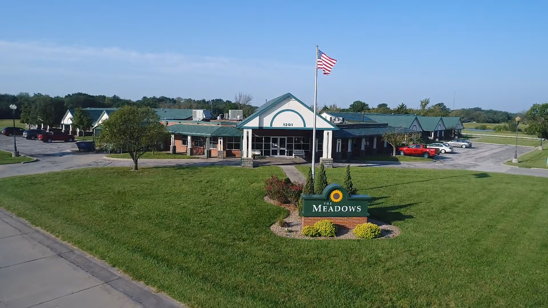 Exterior view of The Meadows facility with a green lawn, sign, flagpole, and parking.