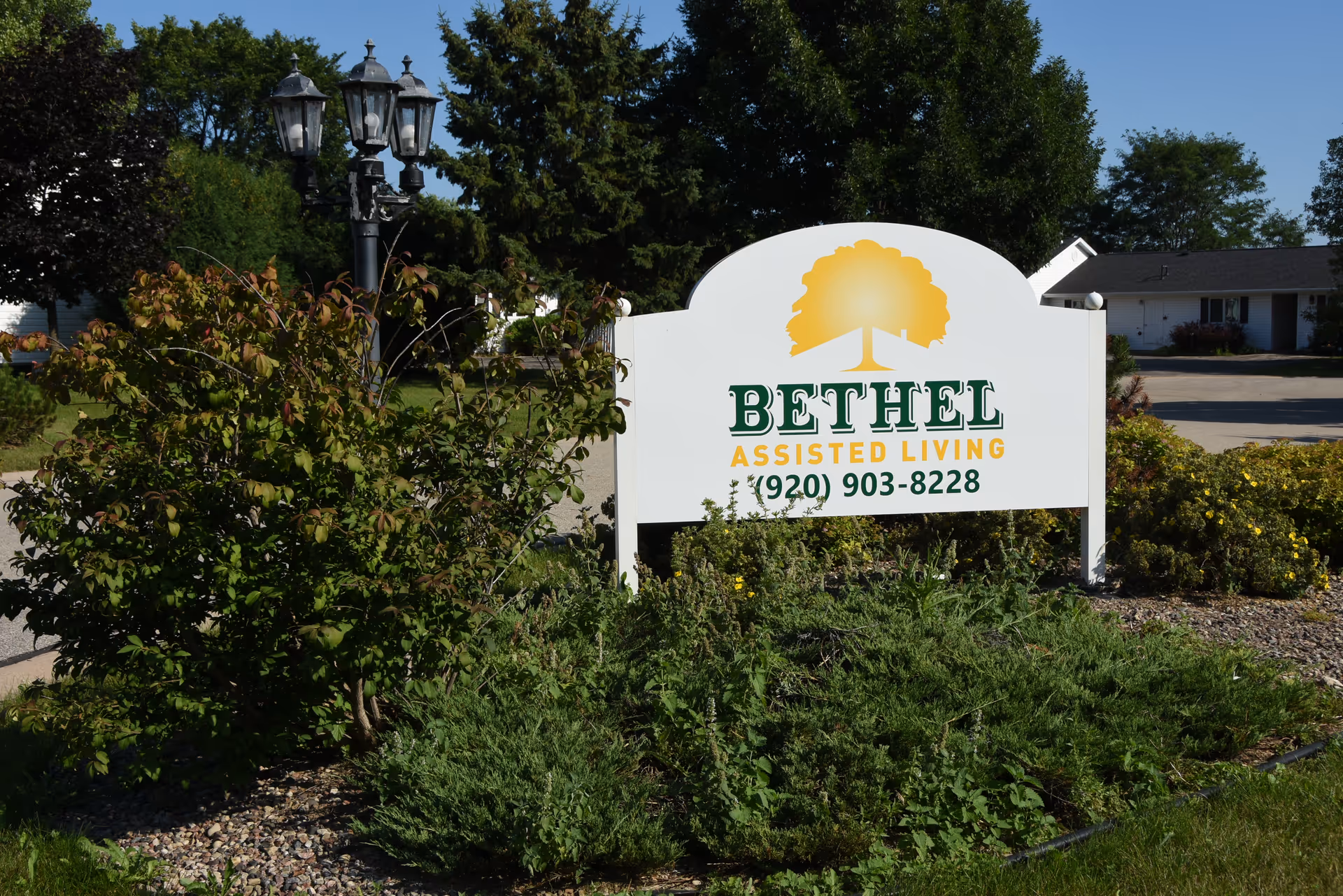 Bethel Assisted Living sign atop a landscaped lawn with trees and a distant building.