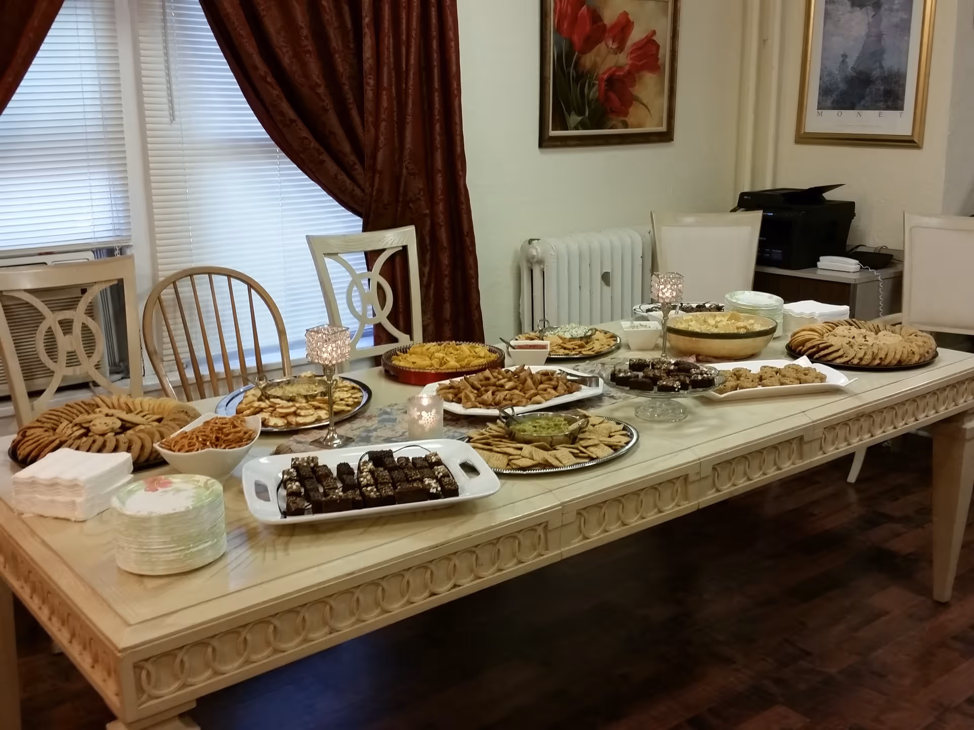 A dining table set with a variety of snacks and desserts including cookies, brownies, pretzels, crackers with dips, and other finger foods. The table is in a room with wooden flooring, cream-colored chairs, red curtains, and framed artwork on the walls.
