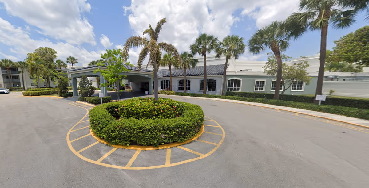 Exterior view of a single-story building with a covered entrance driveway, surrounded by palm trees and neatly trimmed bushes under a partly cloudy sky.