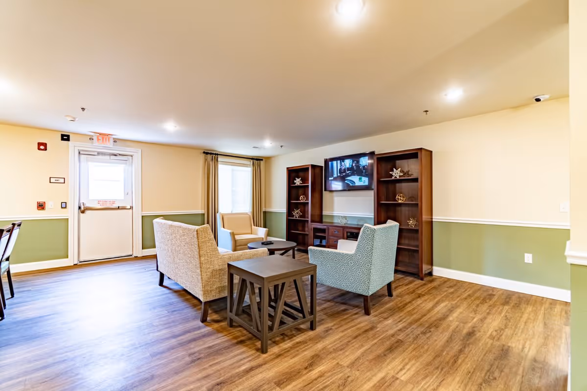 A cozy living room area in a senior living facility with wooden flooring, beige and green walls, and ceiling lights. The room features a beige loveseat, two armchairs in beige and light blue, a small wooden side table, and a round coffee table. There are two wooden bookshelves with decorative items and a flat-screen TV mounted on the wall between them. A window with beige curtains and an exit door are visible in the background.