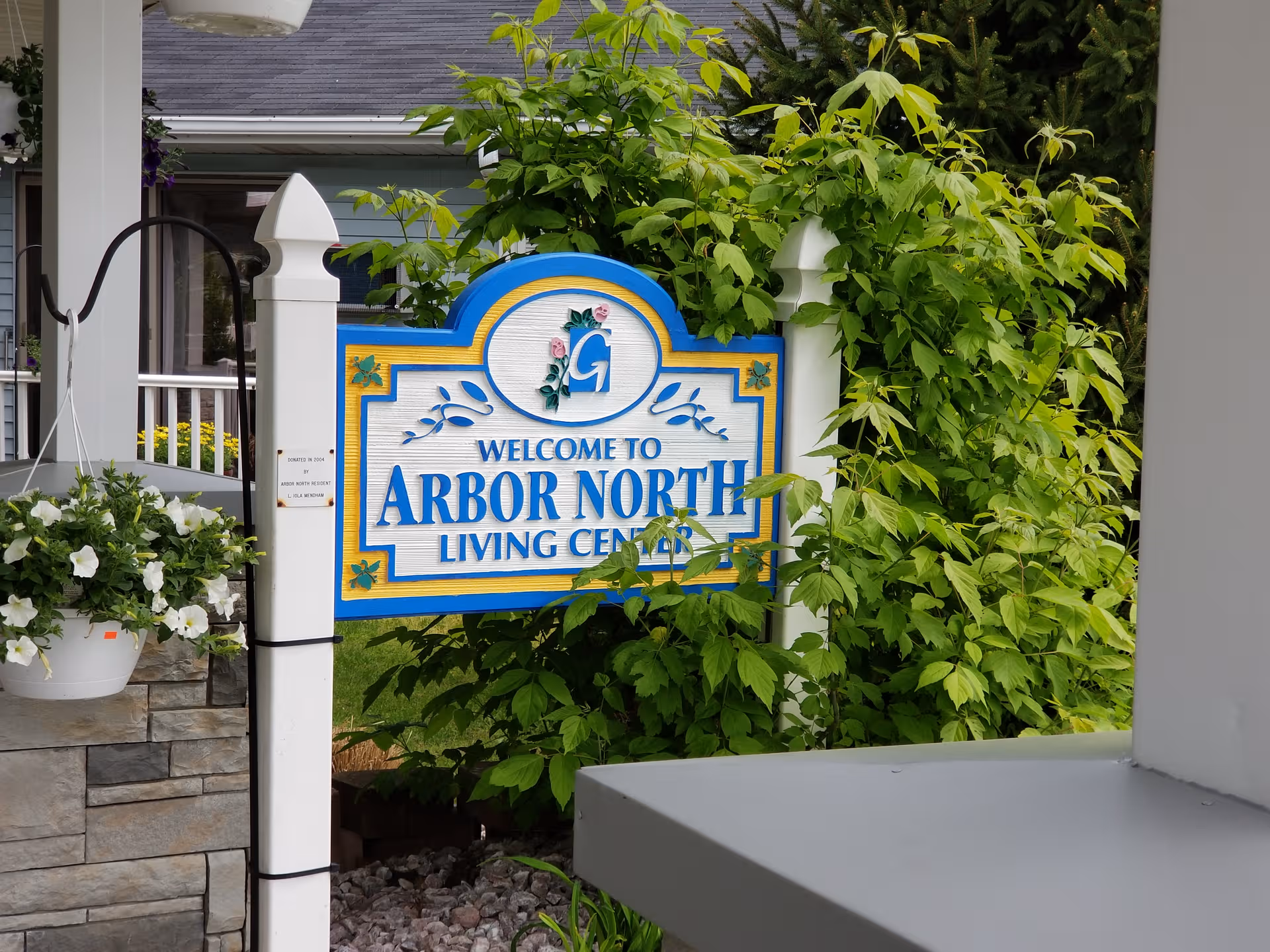 Welcome sign for Arbor North Living Center amid lush greenery on a porch.