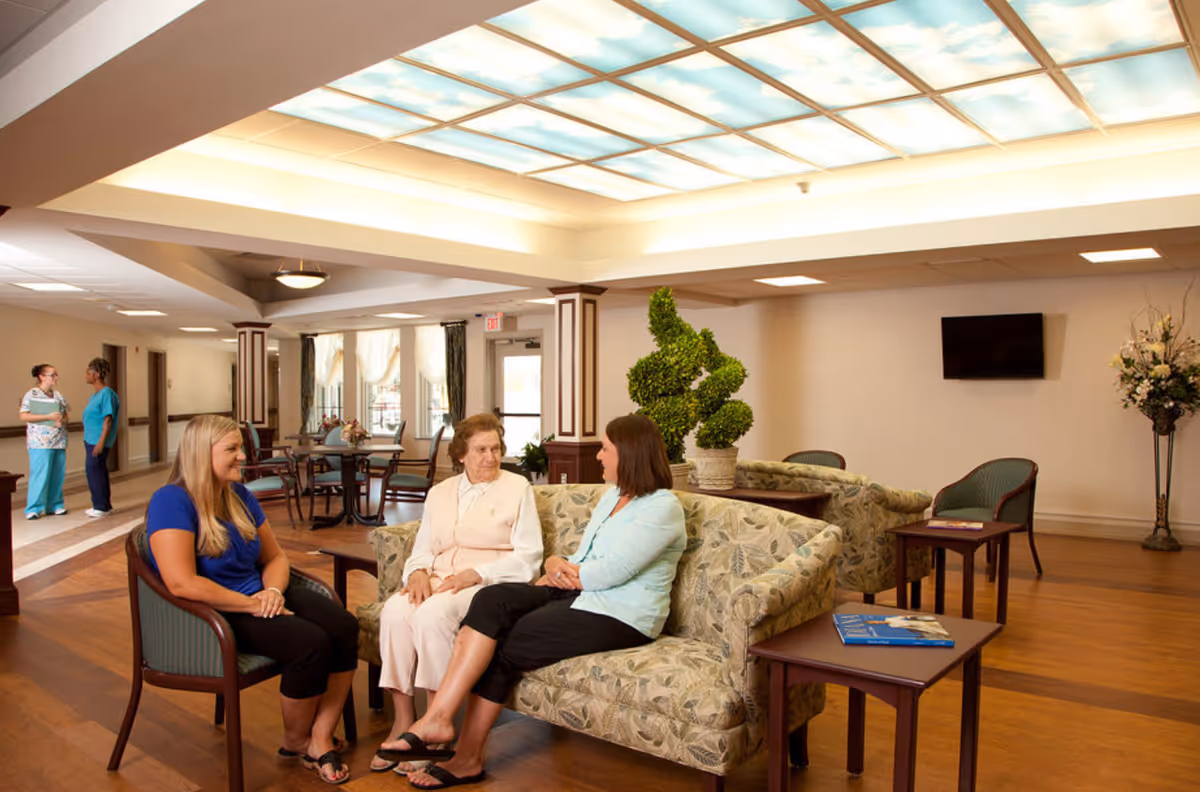 A well-lit common area in a senior living facility with three women sitting and conversing on patterned sofas and chairs. The room features a decorative plant, a wall-mounted TV, wooden flooring, and a ceiling with illuminated panels. In the background, two staff members are standing and talking near a hallway.