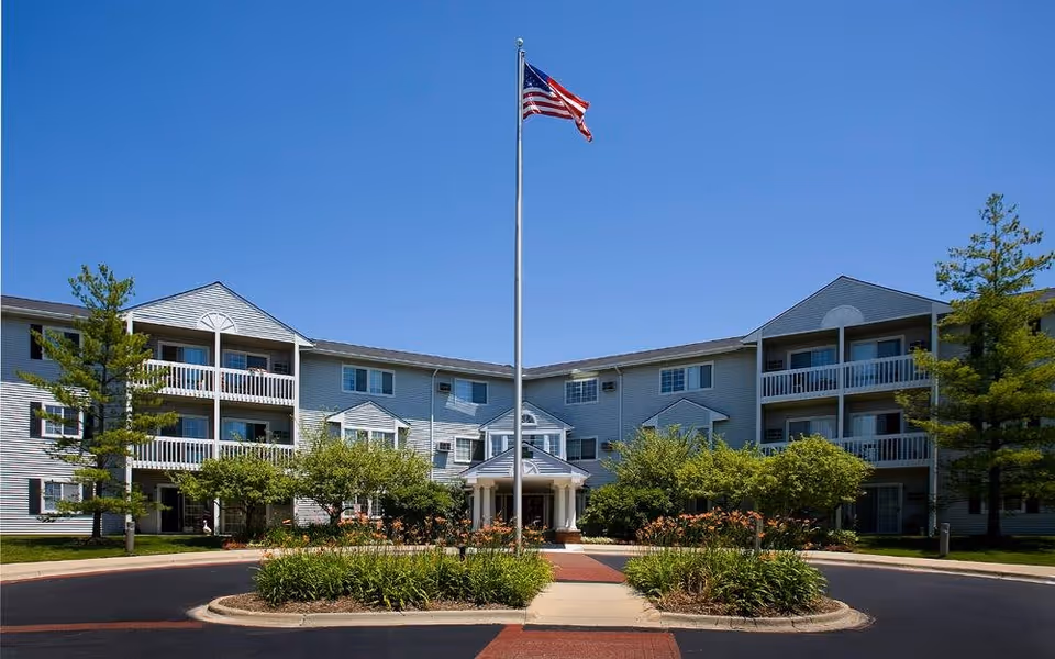 Exterior view of a three-story senior living facility building with balconies, surrounded by trees and landscaping. An American flag flies on a tall flagpole in front of the building under a clear blue sky.