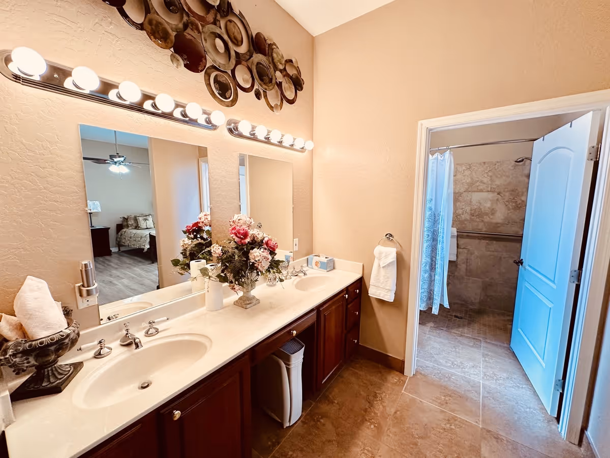 Double-sink bathroom with bright mirrors, flowers, and an adjoining shower.