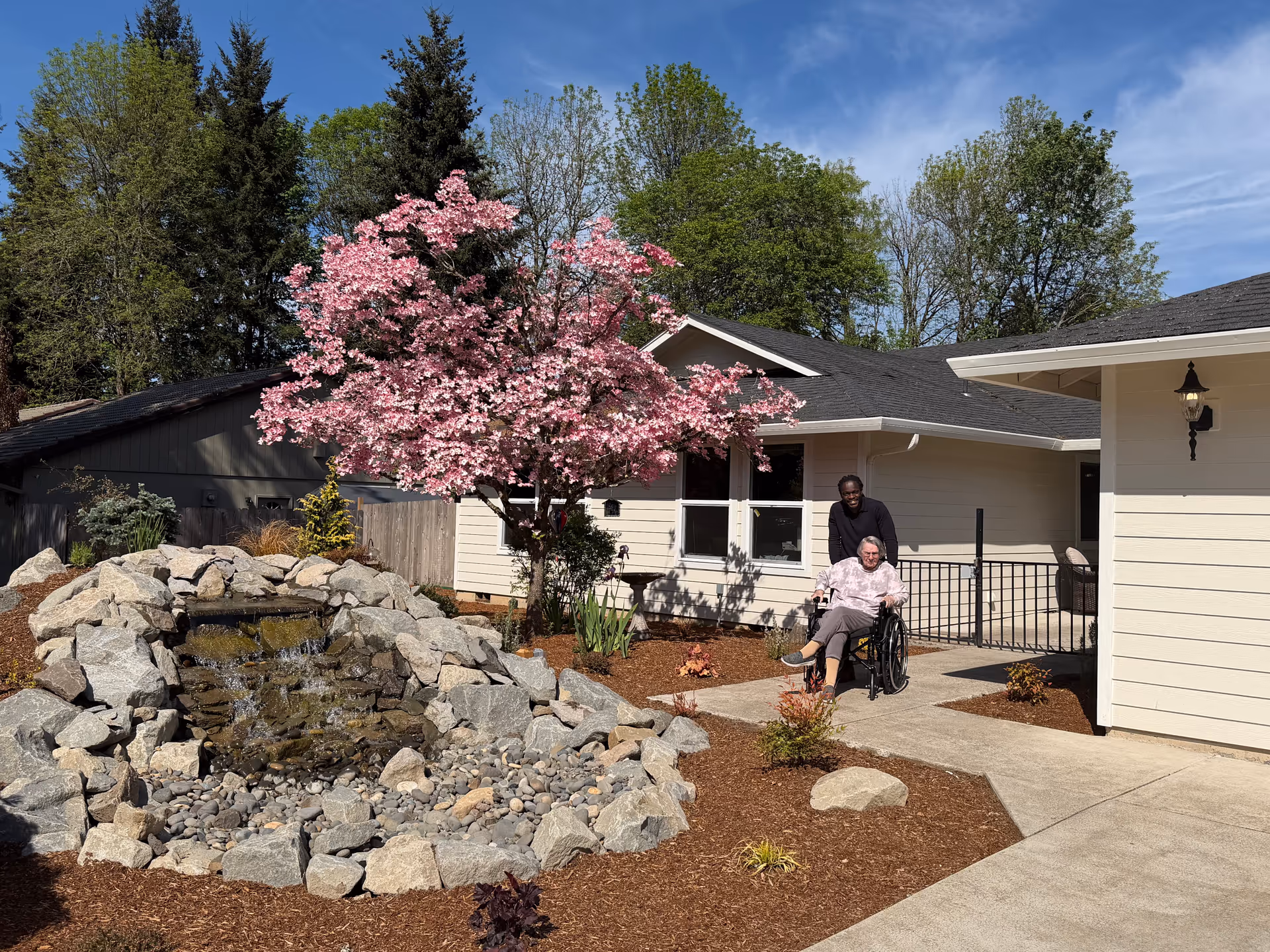 Elderly woman in wheelchair with caregiver outside a single-story care home, surrounded by a flowering tree.