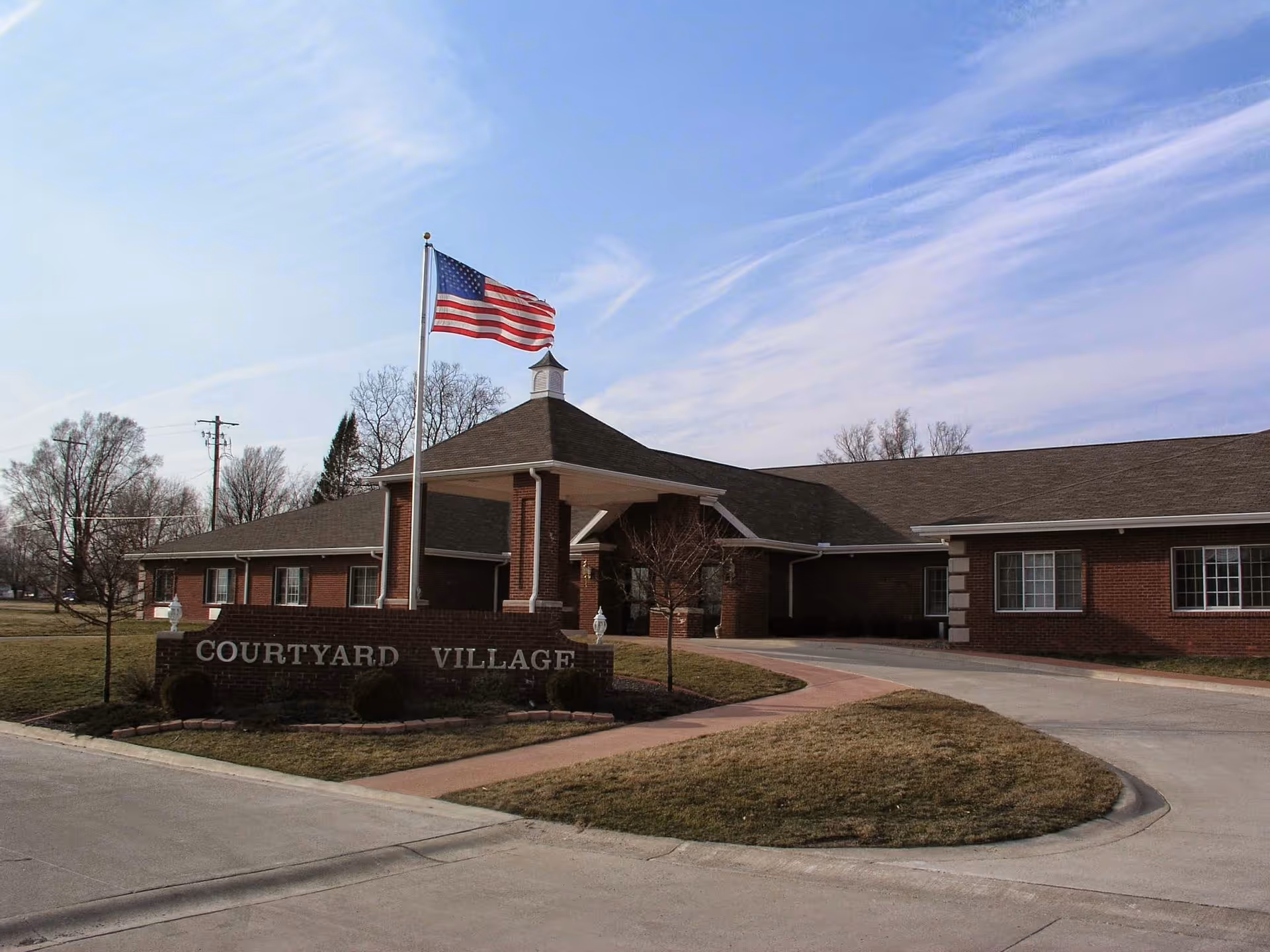 Exterior view of a single-story brick building with a covered entrance and an American flag flying on a flagpole. A sign in front reads 'Courtyard Village'. The sky is partly cloudy and there is a driveway and some grass around the building.