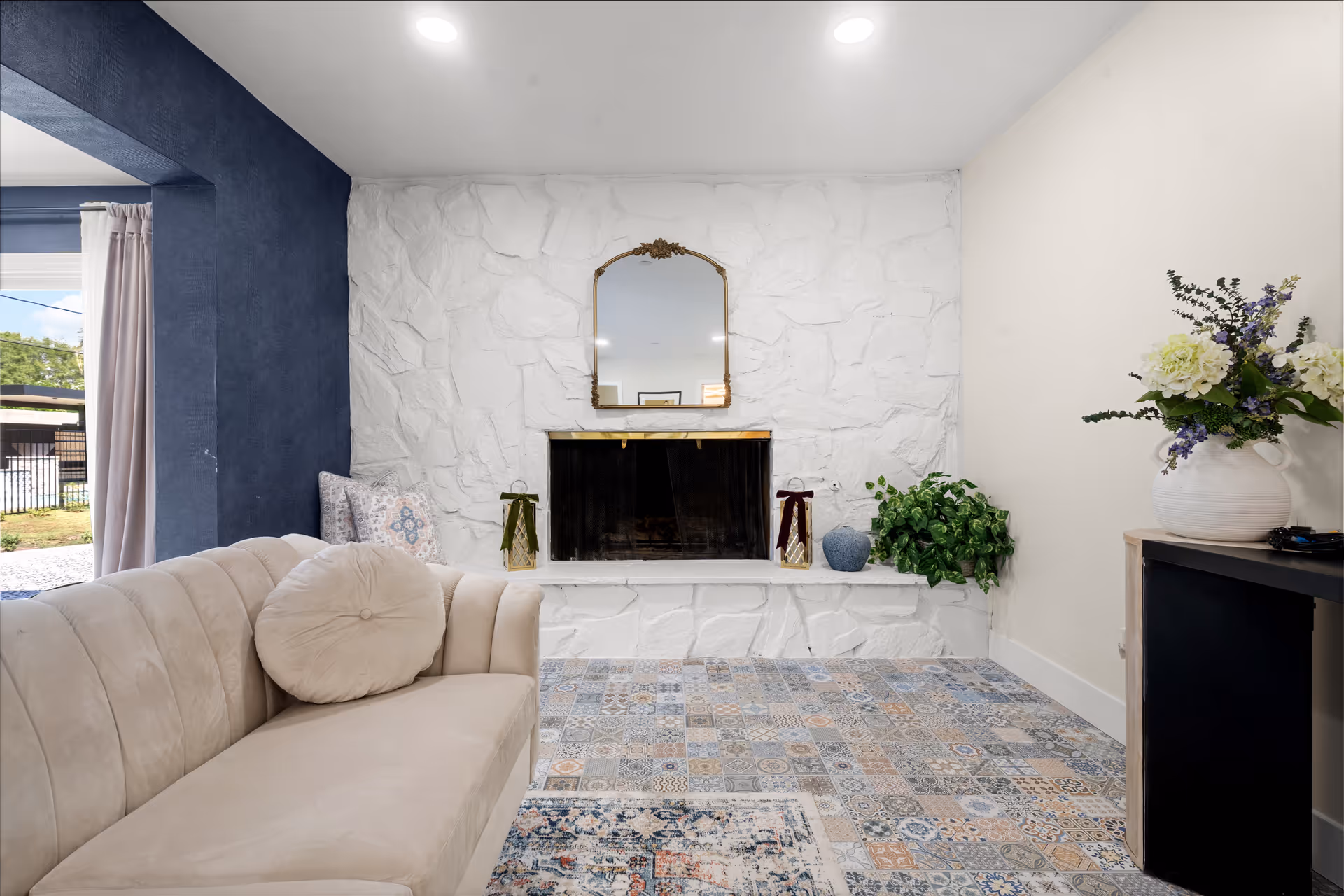 Cozy living room with a stone fireplace, cream sofa, patterned rug, and potted plants.