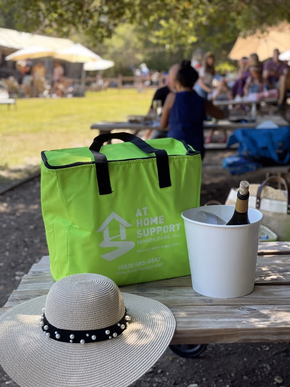 Bright green At Home Support tote on a picnic table with hat and champagne bucket.