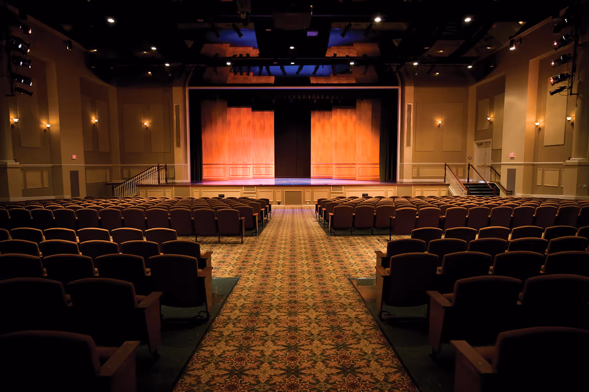 Interior view of an empty auditorium with rows of cushioned seats facing a wooden stage with closed curtains. The room is warmly lit with wall sconces and ceiling lights, and has a patterned carpeted aisle leading to the stage.