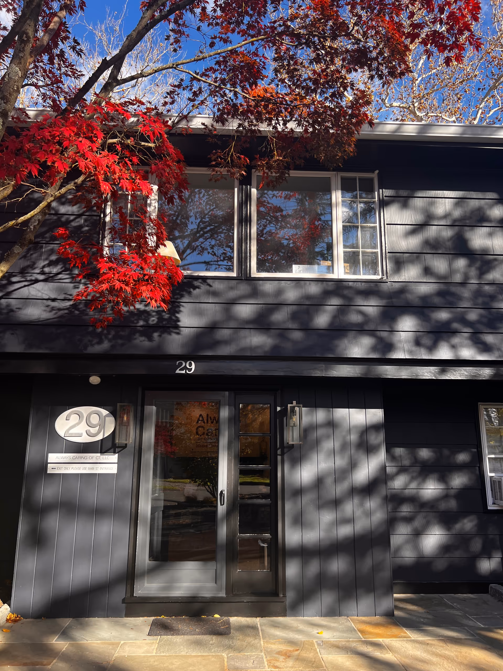 Two-story dark gray building exterior with large windows, a glass door, and a red-leafed tree.
