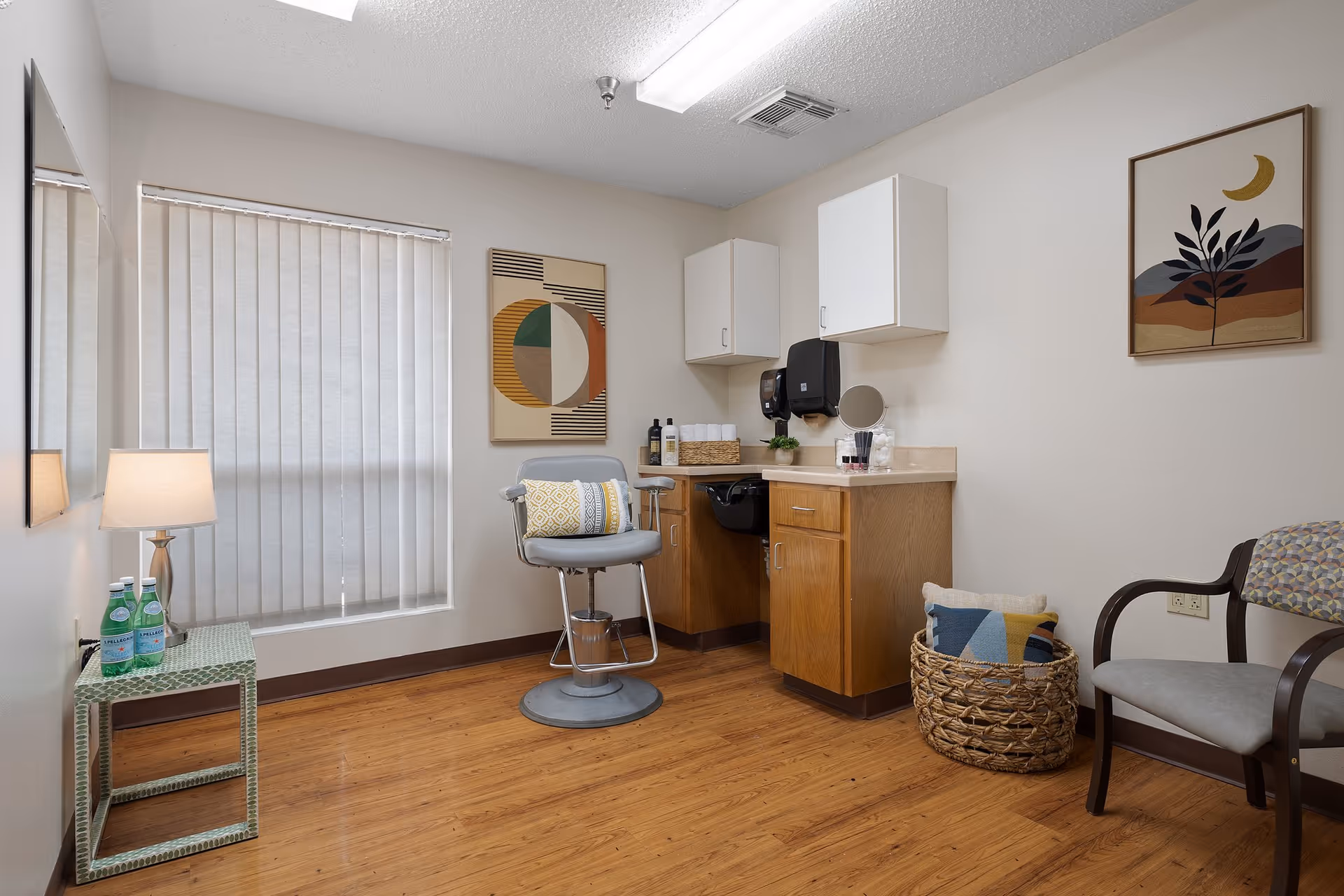 A small, clean room with wooden flooring and white walls featuring a window with vertical blinds. There is a salon-style chair with a decorative pillow in front of a counter with cabinets above and below. The counter holds a basket with towels, bottles, a soap dispenser, and a small mirror. To the right is a woven basket with pillows and a cushioned chair with wooden armrests. On the left side, there is a small table with two bottles of sparkling water and a lamp. Two abstract art pieces hang on the walls.