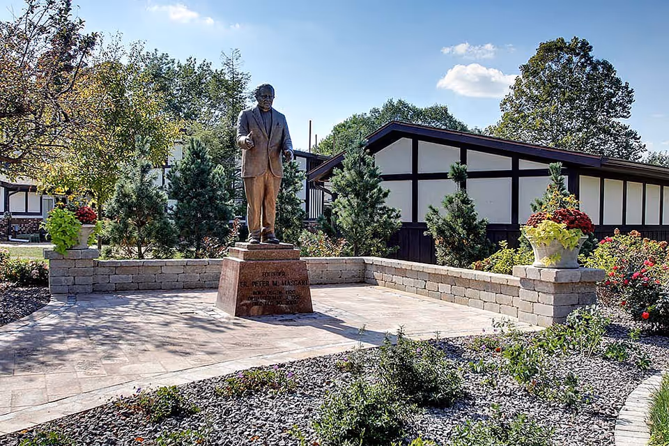 Bronze statue of a man on a pedestal in a landscaped courtyard in front of a single-story building with Tudor-style trim.