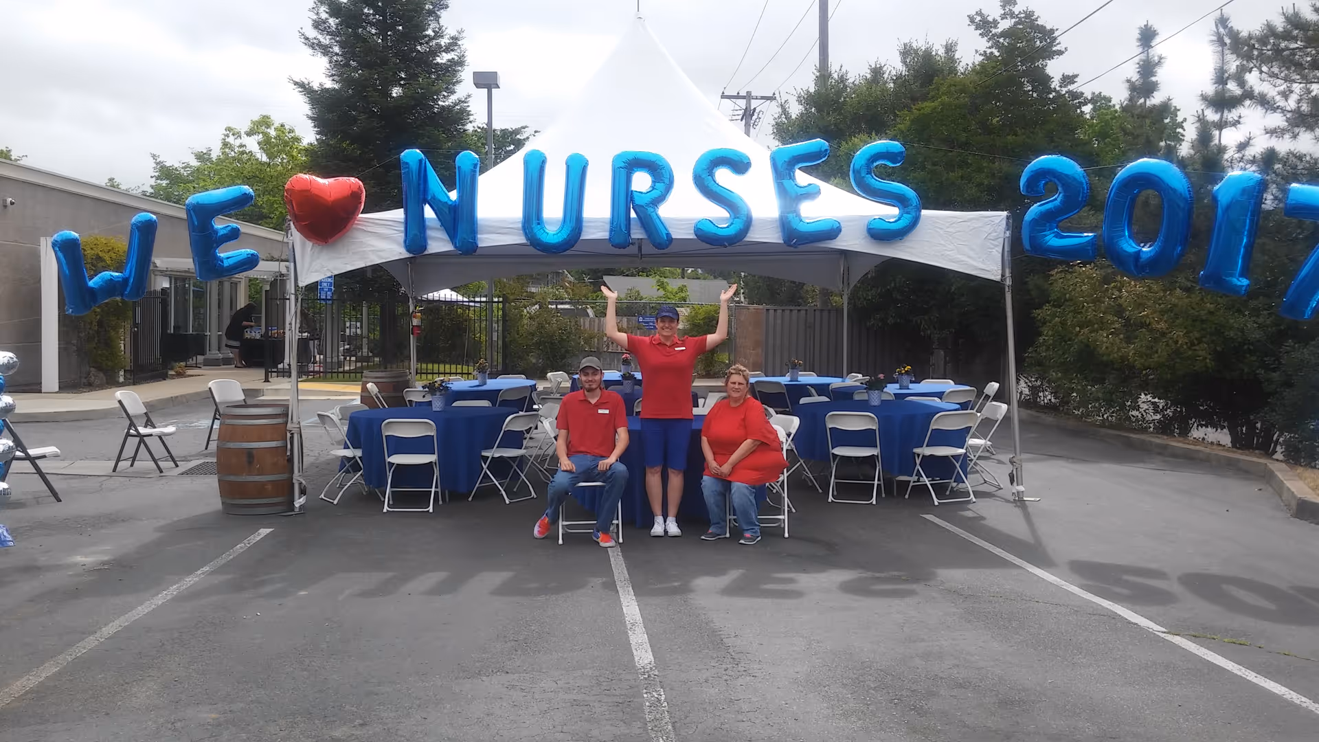 Outdoor event under a white tent with blue balloon letters spelling NURSES and volunteers posing.
