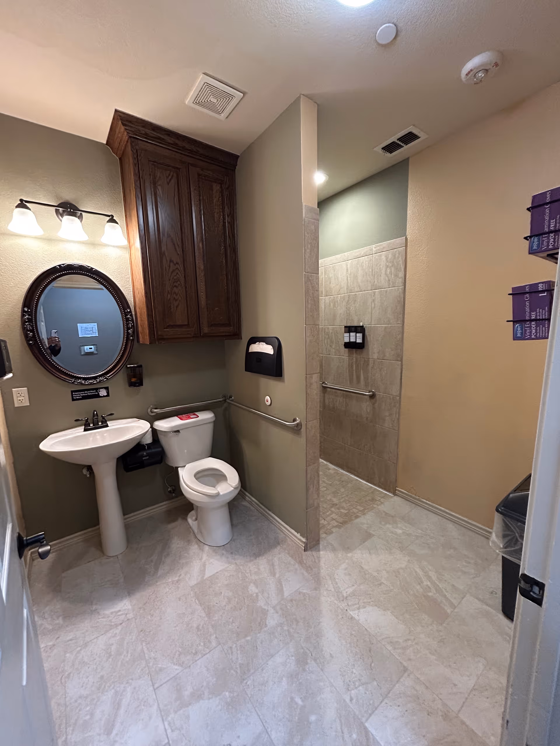 Accessible facility bathroom with pedestal sink, toilet, mirror, cabinet, and tiled shower area.