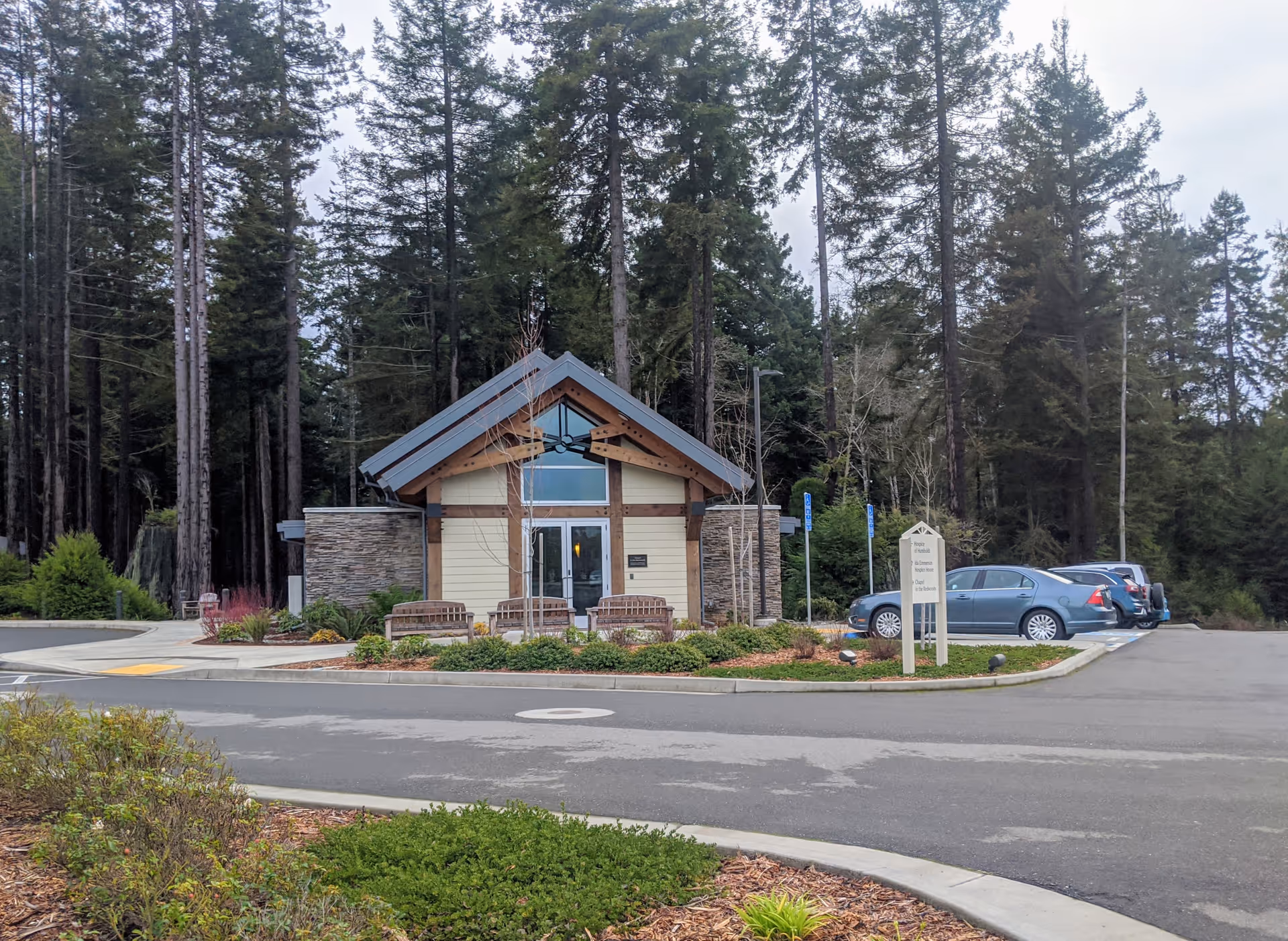 Lodge-style hospice entrance with benches, parking, and a forested backdrop.
