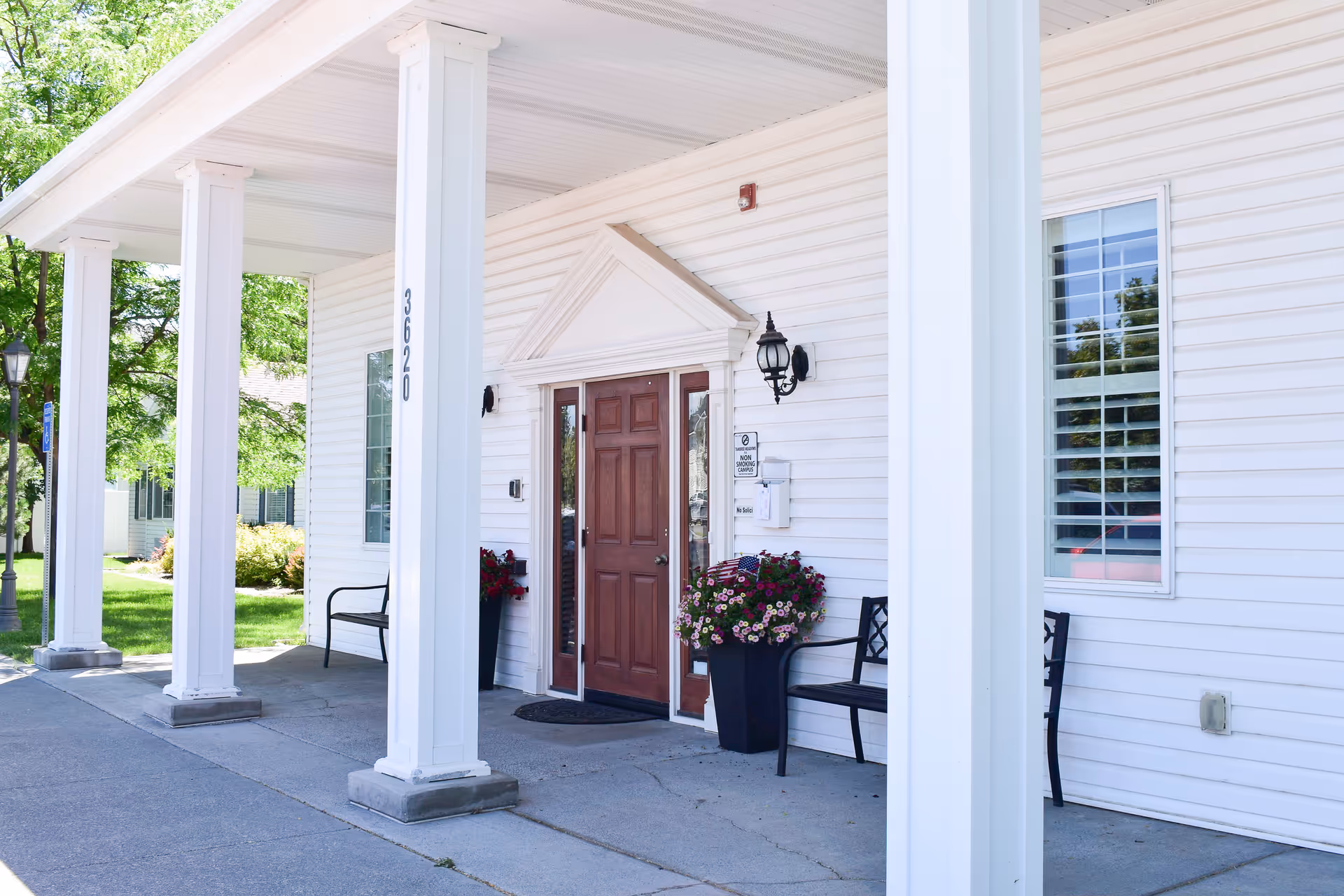 White exterior entrance with wooden door, tall columns, benches, and flower planters.