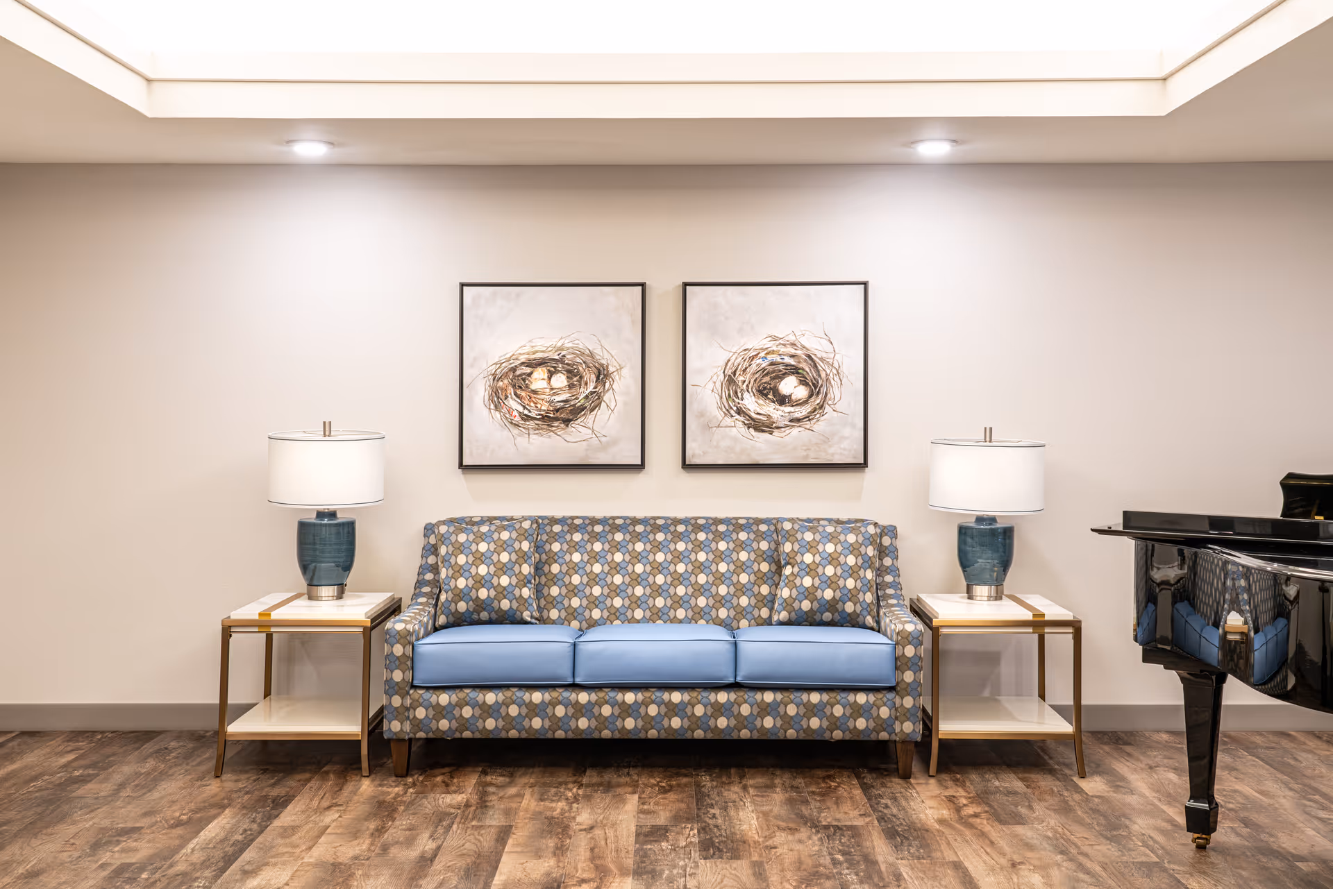 Patterned blue sofa with two side tables, lamps, and a piano in a bright lobby.